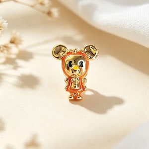 May include: A gold-toned metal charm shaped like a stylised mouse character. The figure features a glossy orange enamel border around its face and body, with black eyes and a small yellow nose. The metal surface is reflective and polished throughout.