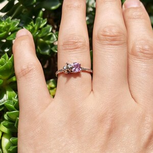 Puede incluir: Un anillo de oro rosa con una piedra preciosa cuadrada de color p&uacute;rpura claro. El anillo presenta peque&ntilde;os detalles decorativos a ambos lados de la piedra preciosa y a lo largo de la banda. El anillo se muestra sobre un fondo de follaje verde.