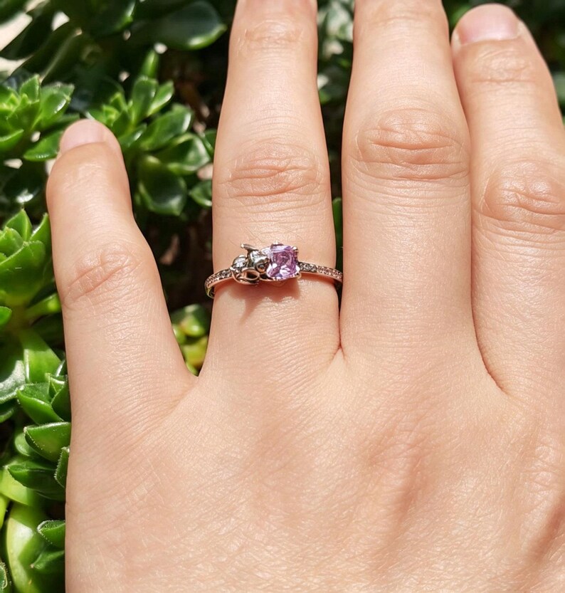 Puede incluir: Un anillo de oro rosa con una piedra preciosa cuadrada de color p&uacute;rpura claro. El anillo presenta peque&ntilde;os detalles decorativos a ambos lados de la piedra preciosa y a lo largo de la banda. El anillo se muestra sobre un fondo de follaje verde.