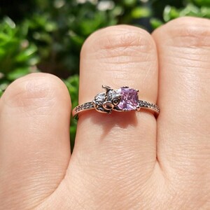 Puede incluir: Un anillo plateado con un dise&ntilde;o de cerdo y una gema cuadrada de color p&uacute;rpura claro. La banda del anillo est&aacute; adornada con peque&ntilde;as piedras transparentes. La cara del cerdo est&aacute; detallada con ojos y hocico.