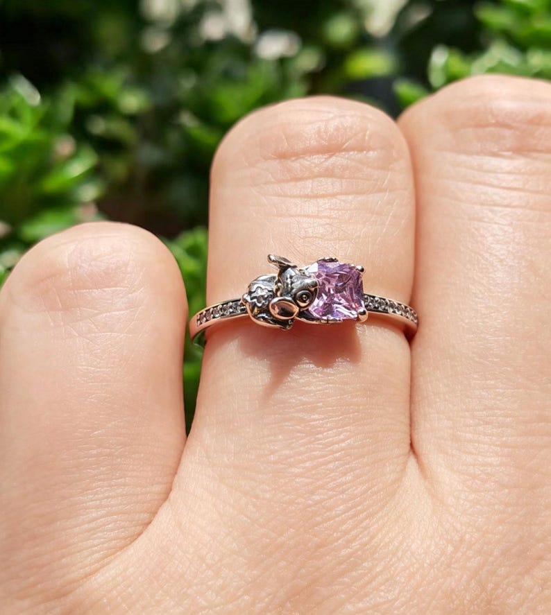 Puede incluir: Un anillo plateado con un dise&ntilde;o de cerdo y una gema cuadrada de color p&uacute;rpura claro. La banda del anillo est&aacute; adornada con peque&ntilde;as piedras transparentes. La cara del cerdo est&aacute; detallada con ojos y hocico.