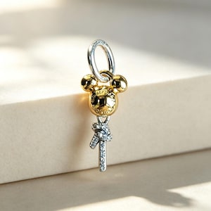 Könnte beinhalten: Ein goldfarbener Anhänger in Form eines kugelförmigen Mickey-Maus-Kopfes mit eingraviertem Disney-Logo auf der Vorderseite. Darunter befindet sich ein silberfarbenes, T-förmiges Element mit kleinen klaren Steinen, befestigt an einem silbernen Ring.