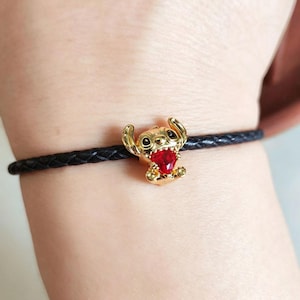 Peut inclure: Bracelet avec un charm dor&eacute; repr&eacute;sentant Stitch, orn&eacute; d'un c&oelig;ur rouge. Le charm est fix&eacute; &agrave; un bracelet en cuir tress&eacute; noir. Accessoire sur le th&egrave;me de Disney.