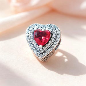 Puede incluir: Un charm en forma de corazón con una piedra roja facetada central rodeada por dos filas concéntricas de cristales transparentes engastados en pavé. El marco de metal es plateado con un patrón recortado decorativo en los bordes laterales.