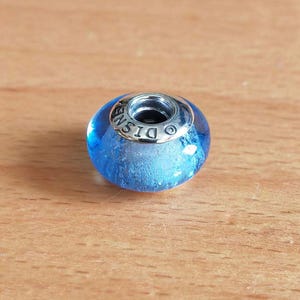 Peut inclure: Une perle de verre bleue avec un cœur en métal argenté. La perle a un intérieur givré et le mot "DISNEY" est gravé sur le cœur en métal. La perle est ronde et a une surface lisse.
