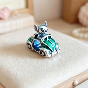 May include: A silver-toned charm featuring a cartoon character driving a green and blue car. The charm has a detailed design with a glossy finish. The car has silver wheels and a blue interior. The character is silver and blue.