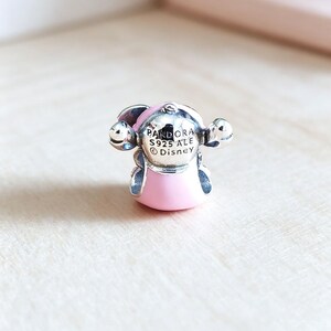 Pu&ograve; includere: Un charm Pandora argentato con un design a personaggio rosa e argento. Il charm ha un corpo e una testa rosa, con dettagli argentati e la scritta "PANDORA S925 ALE Disney". Il charm &egrave; su una superficie di legno chiaro.