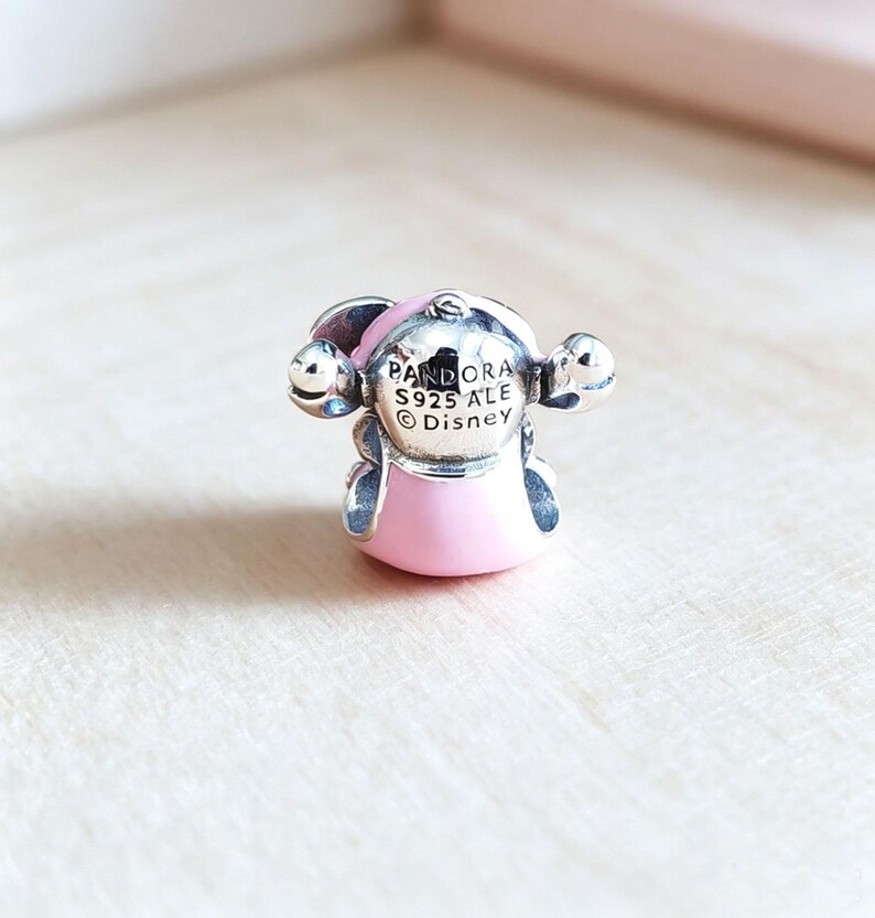 Pu&ograve; includere: Un charm Pandora argentato con un design a personaggio rosa e argento. Il charm ha un corpo e una testa rosa, con dettagli argentati e la scritta "PANDORA S925 ALE Disney". Il charm &egrave; su una superficie di legno chiaro.