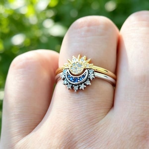 May include: A two-tone ring set featuring a gold band with a sunburst design adorned with clear stones and a silver band with a crescent moon motif set with blue stones, accented by small star-shaped details and clear stones along the edge.