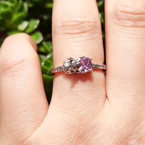Puede incluir: Un anillo de oro rosa con una gema cuadrada de color p&uacute;rpura claro. El anillo tiene un peque&ntilde;o dise&ntilde;o detallado en un lado y peque&ntilde;as piedras negras a lo largo de la banda. El anillo se muestra en un dedo.