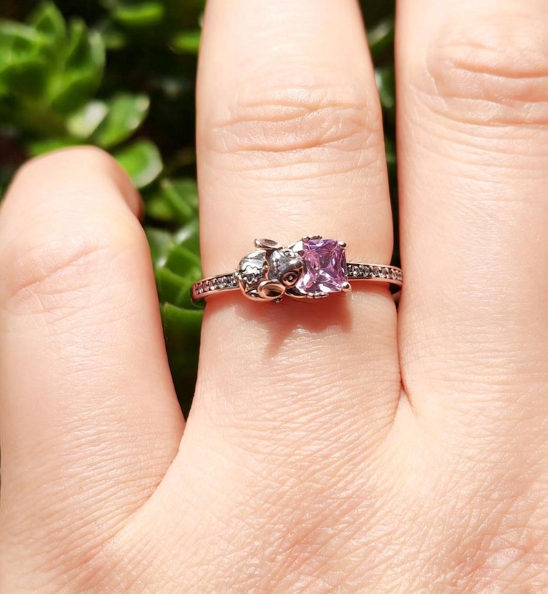 Puede incluir: Un anillo de oro rosa con una gema cuadrada de color p&uacute;rpura claro. El anillo tiene un peque&ntilde;o dise&ntilde;o detallado en un lado y peque&ntilde;as piedras negras a lo largo de la banda. El anillo se muestra en un dedo.