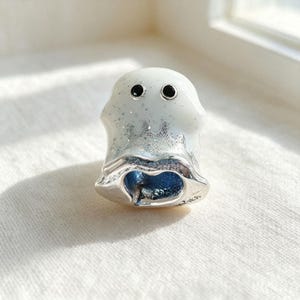 Op de afbeelding: Een zilveren bedel in de vorm van een geest met een witte emaille toplaag voorzien van subtiele glitteraccenten. De geest heeft twee kleine zwarte ronde ogen en een holle opening aan de onderkant, met een zilverkleurige afwerking onderaan.