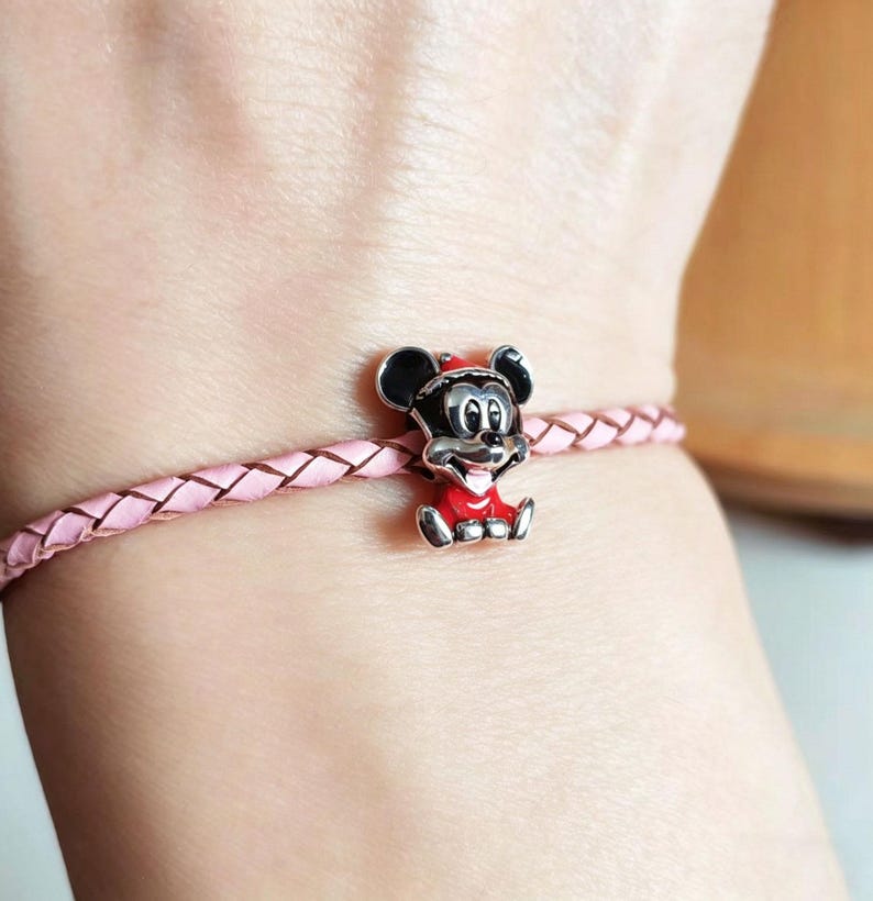 K&ouml;nnte beinhalten: Ein rosa geflochtenes Lederarmband mit einem silbernen Mickey Mouse-Anh&auml;nger. Der Mickey Mouse-Anh&auml;nger tr&auml;gt ein rotes Outfit und einen roten Hut. Das Armband wird am Handgelenk getragen.