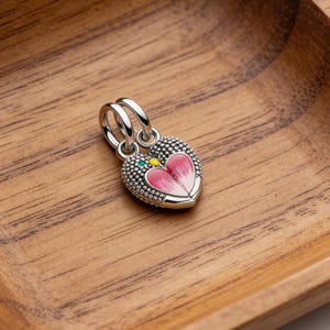 May include: A silver heart-shaped charm with a pink and white floral design. The charm has a textured surface and is accented with green and yellow details. It features two loops for attachment.