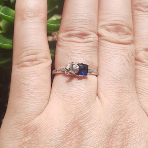 May include: A silver ring with a dark blue, square-cut gemstone. The ring features a decorative design on one side and small, clear stones along the band. The ring is shown on a hand.