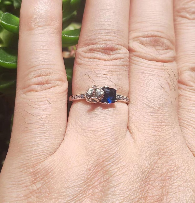 May include: A silver ring with a dark blue, square-cut gemstone. The ring features a decorative design on one side and small, clear stones along the band. The ring is shown on a hand.