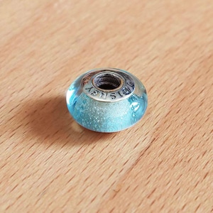 Peut inclure: Une perle ronde bleue et argentée avec le mot "DISNEY" gravé sur la bande argentée. La perle a une couche extérieure transparente bleu clair et une couche intérieure scintillante bleu clair. La perle est sur une surface en bois.