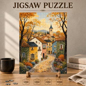 May include: A jigsaw puzzle featuring a painting of a European village street with stone buildings, autumn trees, and a church tower. The puzzle is displayed on a wooden stand with options for 200, 300, 500, or 1000 pieces printed at the bottom.
