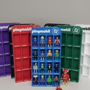 Op de afbeelding: Een verzameling Playmobil DC Comics-figuren in kleurrijke, verticale display-rekken. De rekken zijn paars, zwart, rood, blauw, groen en wit. Het blauwe rek bevat de figuren, de andere zijn leeg.