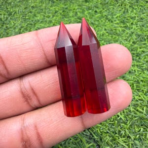 May include: Two polished, deep red, hexagonal crystal points with smooth, faceted surfaces and pointed tips. These translucent, cylindrical stone towers exhibit a vibrant crimson hue and a clear, glass-like texture, standing side by side.