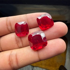 Puede incluir: Tres piedras preciosas de color rojo vibrante, talladas en forma de cojín con superficies facetadas. Las piedras son translúcidas y muestran un tono carmesí profundo, reflejando la luz en sus bordes pulidos. Estas gemas cuadradas están agrupadas.