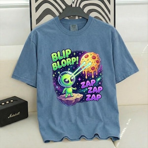 May include: Blue short-sleeved t-shirt featuring a green alien character zapping a pizza with the phrases "BLIP BLORP!" and "ZAP ZAP ZAP". The alien is on a floating rock against a purple and blue galaxy backdrop.