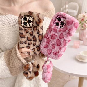 May include: Two fluffy phone cases with leopard print designs. One case is brown and tan, the other is pink. Both cases have a matching pom-pom keychain with pearl accents. The cases are designed to protect the phone while adding a stylish flair.