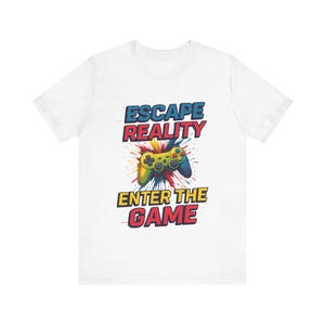 May include: White t-shirt with a vibrant graphic. It shows a video game controller and the text "ESCAPE REALITY ENTER THE GAME" in blue, red, and yellow. The design includes a splash of colours.