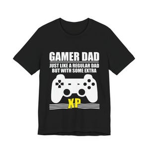 Op de afbeelding: Zwart t-shirt met de tekst "GAMER DAD" boven een witte controller-afbeelding. De tekst "JUST LIKE A REGULAR DAD BUT WITH SOME EXTRA" staat eronder. Het woord "XP" staat in het geel onder de controller.