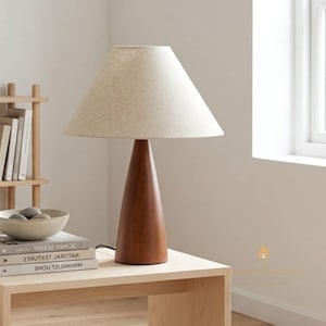Pode incluir: Candeeiro de mesa em madeira com base cónica castanha escura e abajur creme. O candeeiro está numa mesa de madeira clara, com livros e uma tigela decorativa. Design minimalista e moderno, ideal para casa.