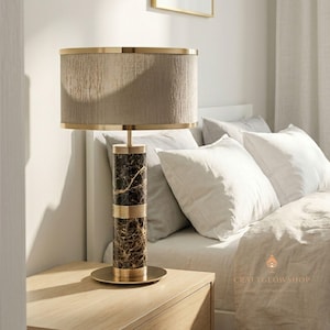Puede incluir: Una lámpara de mesa decorativa con una pantalla beige y un marco de metal dorado. La base de la lámpara es un cilindro de mármol oscuro con detalles dorados. La lámpara está sobre una mesita de noche de madera.