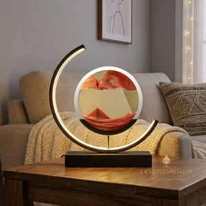 Puede incluir: Una moderna lámpara de arte de arena con una pantalla de vidrio circular que contiene arena roja y beige. La lámpara tiene una base negra y un marco curvo de color dorado con iluminación integrada. La lámpara está sobre una mesa de madera.