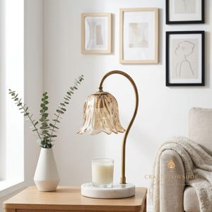 May include: A decorative table lamp with a flower-shaped glass shade in shades of amber and gold. The lamp has a curved brass stem and sits on a marble base. A white candle is on the base. A white vase with green stems is on the left.
