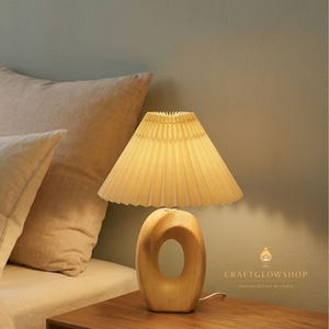May include: A table lamp featuring a light-coloured wooden base with a central oval cutout and a pleated, off-white conical lampshade. The lamp emits a warm glow and includes the text CRAFTGLOWSHOP SPECIALIZING IN LAMPS printed on the lower right.