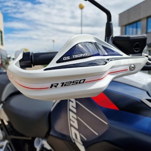 May include: White and blue motorcycle handguards with the text "GS TROPHY" and "R 1250" in black. The handguards have a red stripe and are mounted on a motorcycle with a blue and black colour scheme.
