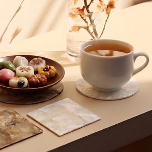 Può includere: Una natura morta con una tazza bianca di tè, un piatto di pasticcini colorati e sottobicchieri decorativi. La scena è immersa in una luce calda, creando un'atmosfera accogliente. È presente anche un vaso con fiori.