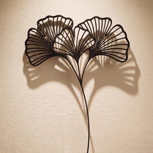 Peut inclure: Décoration murale en métal noir en forme de feuilles de ginkgo. La sculpture présente deux feuilles de ginkgo stylisées avec des détails de veines complexes, montées sur une fine tige. Les feuilles ont une finition texturée et scintillante.