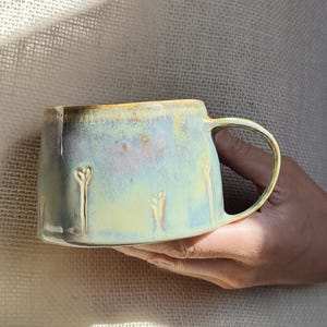 May include: A handcrafted ceramic mug with a distinctive form and a pale blue and yellow glaze. The mug has a curved handle and embossed floral patterns. The mug is held in a person's hand.
