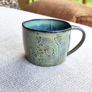 Könnte beinhalten: Handgefertigte Keramik-Tasse mit blau-grüner Glasur. Die Tasse hat geätzte Blumendesigns und einen gebogenen Griff. Das Innere der Tasse ist leuchtend blau. Geeignet für Heißgetränke.