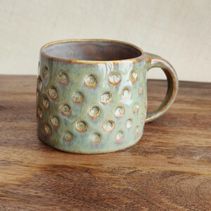 May include: A handmade ceramic mug with a distinctive textured surface. The mug is a muted green colour with a speckled pattern and a brown rim and handle. The mug is on a wooden surface.