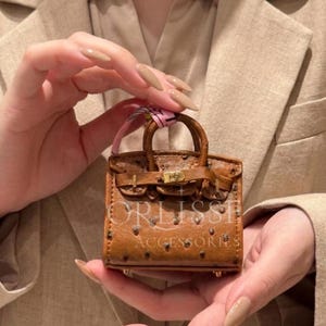 May include: A miniature brown handbag with a textured ostrich-like pattern, featuring two top handles, a gold-tone metal clasp closure, and a small pink ribbon accent. The text ORLISSE ACCESSORIES is printed in white across the front of the bag.