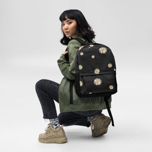 Könnte beinhalten: Ein schwarzer Rucksack mit einem sich wiederholenden Muster aus weißen und gelben Gänseblümchen. Die Tasche verfügt über ein Hauptfach und eine kleinere Fronttasche mit Reißverschluss sowie verstellbare schwarze Schultergurte.