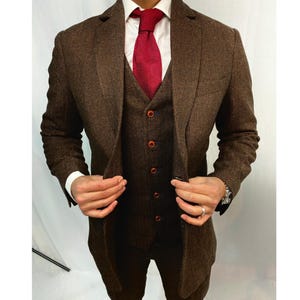 Könnte beinhalten: Ein dreiteiliger brauner Tweedanzug mit roter Krawatte und weißem Hemd. Der Anzug besteht aus einem Blazer, einer Weste und einer Hose. Die Weste hat rote Knöpfe. Der Anzug ist ein klassischer Stil.