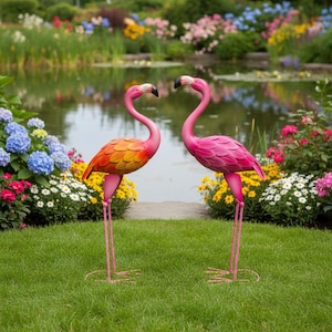 Puede incluir: Dos estatuas de flamencos rosas de jardín, con largas patas y cuellos curvos, se alzan sobre un césped verde. Un flamenco tiene plumas naranjas y amarillas, mientras que el otro es de color rosa liso. El fondo presenta un estanque y flores coloridas.