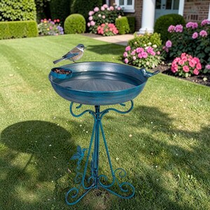 K&ouml;nnte beinhalten: Ein teal-farbenes Vogelbad aus Metall mit einer kleinen, befestigten Schale und einem dekorativen Vogel am Rand. Das Vogelbad wird von einem dreibeinigen St&auml;nder mit Schn&ouml;rkeln getragen. Ein kleiner Vogel sitzt auf dem Rand des Vogelbades.