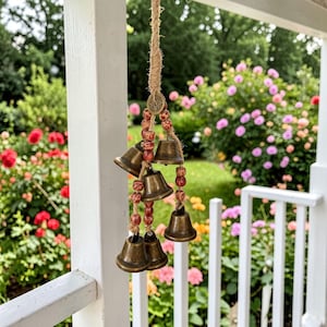 May include: A hanging wind chime featuring six bronze-colored bells suspended from a natural fiber rope. The bells are strung with wooden beads in shades of red and brown. The background shows a garden with colorful flowers.