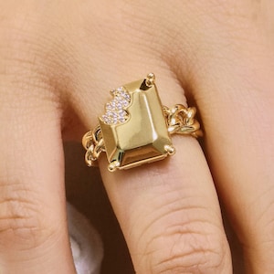 May include: A gold ring with a large, rectangular, emerald-cut central stone. The ring has a chain-link band and a small cluster of sparkling stones. The ring is worn on a finger. The ring is a piece of jewellery.