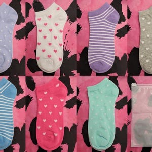 May include: A selection of ankle socks in different colours and designs, such as polka dots, hearts, and stripes. The socks are presented on a pink and black patterned backdrop. A clear plastic bag is also visible.