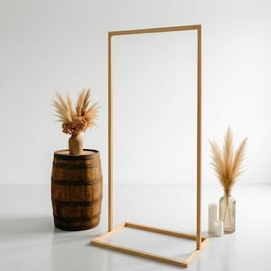 May include: A wooden rectangular frame with a clear center stands on a light-colored floor. A rustic wooden barrel holds a vase of dried flowers. A glass vase with dried grass and two white candles are on the right.