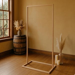 May include: A light-colored wooden rectangular frame, possibly for a backdrop or display, stands on a wooden floor. A wooden barrel with metal bands and a vase of dried flowers are nearby. The room has a warm, inviting atmosphere.